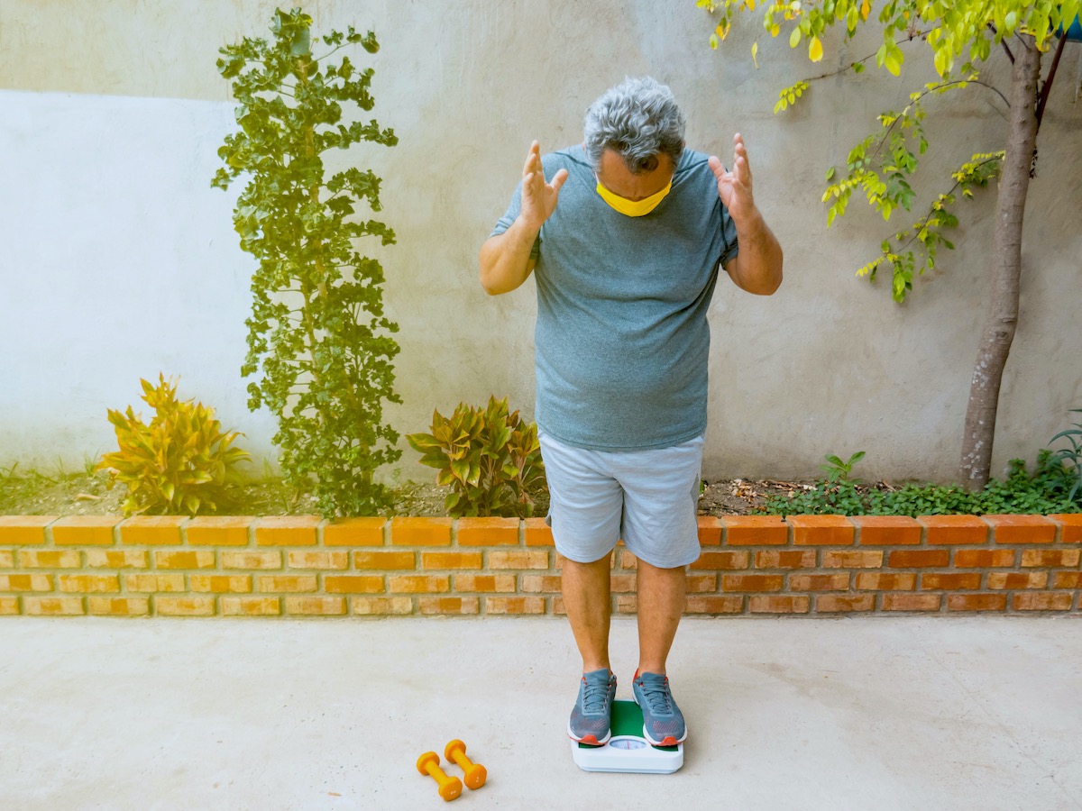 Masked, Overweight Man Standing On A Scale With Arms Raised In Displeasure